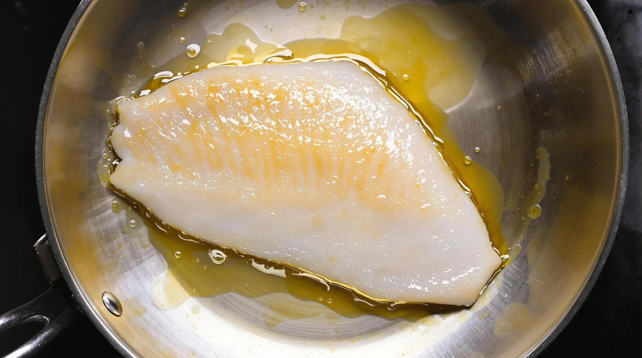 Branzino fillet cooking in stainless steel pan, flesh turning opaque with golden oil bubbling around edges, viewed from above