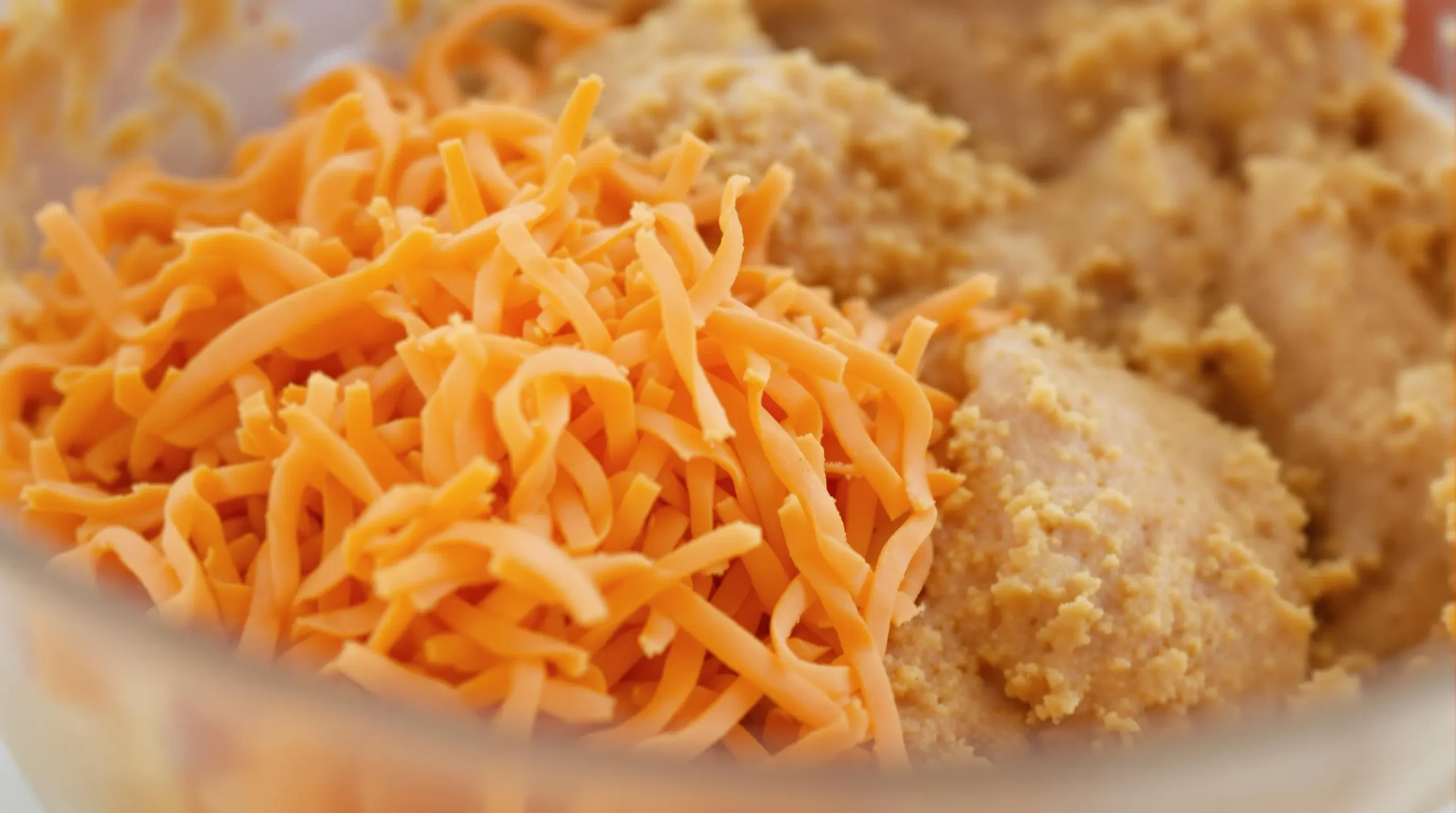 Fresh orange carrots being folded into golden spiced cake batter in glass mixing bowl, showing marbled swirls and texture