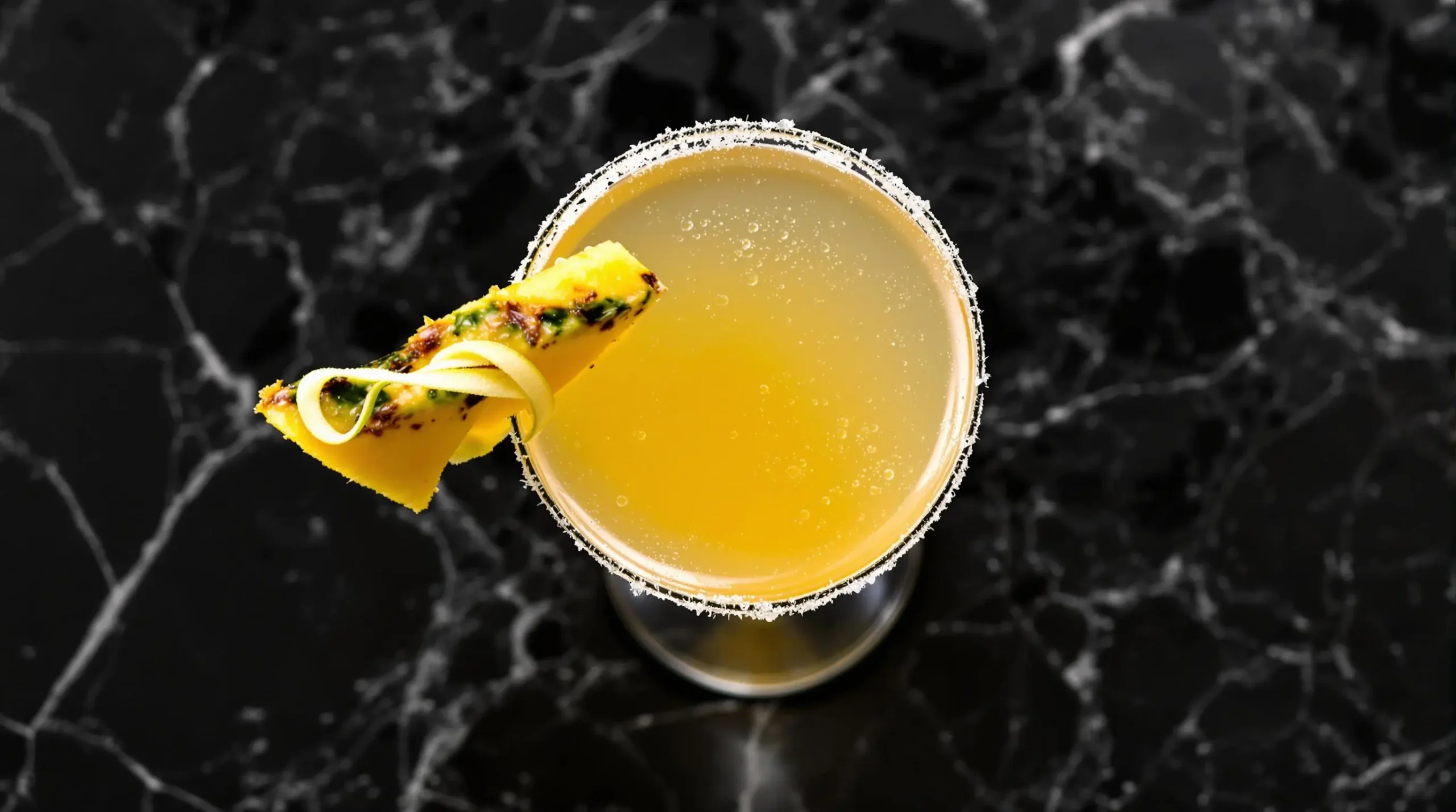 Overhead view of golden pineapple martini with sugar rim, pineapple wedge and lime twist garnish on black stone surface with perfect lighting