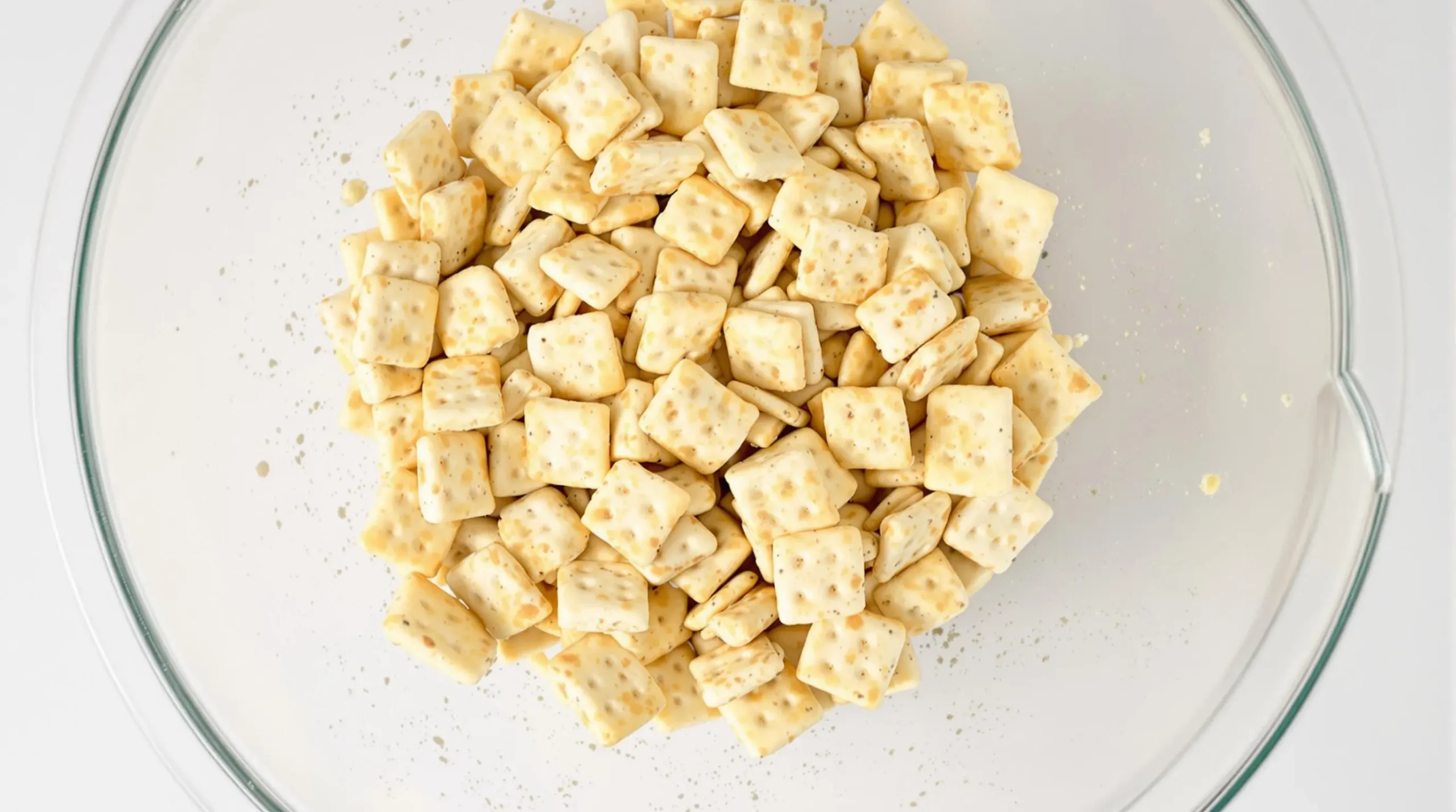 Ranch crackers being tossed in clear glass bowl with seasoning and oil, showing active coating process with visible spice particles